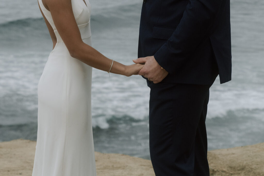 bride and groom holding hands on cliffside as ocean rolls in the background