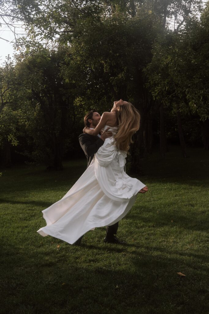 Groom spinning bride around joyfully in the courtyard of an Italian villa, surrounded by sunlit stone walls and vibrant greenery, capturing their carefree wedding day happiness